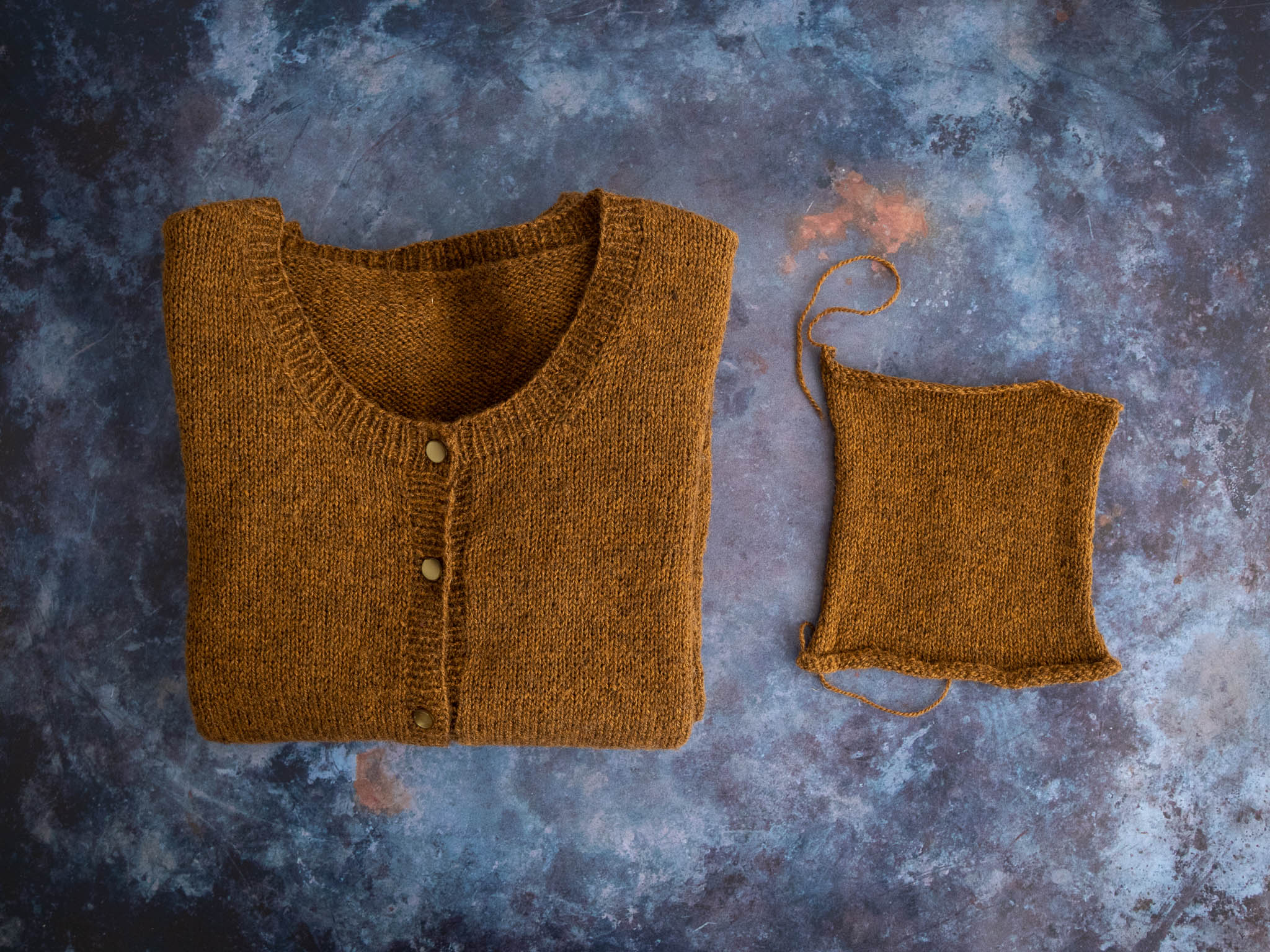 A honey brown cardigan with round neck is folded and lies on a moody dark blue flat surface. To the right of the sweater is a small swatch in the same yarn.
