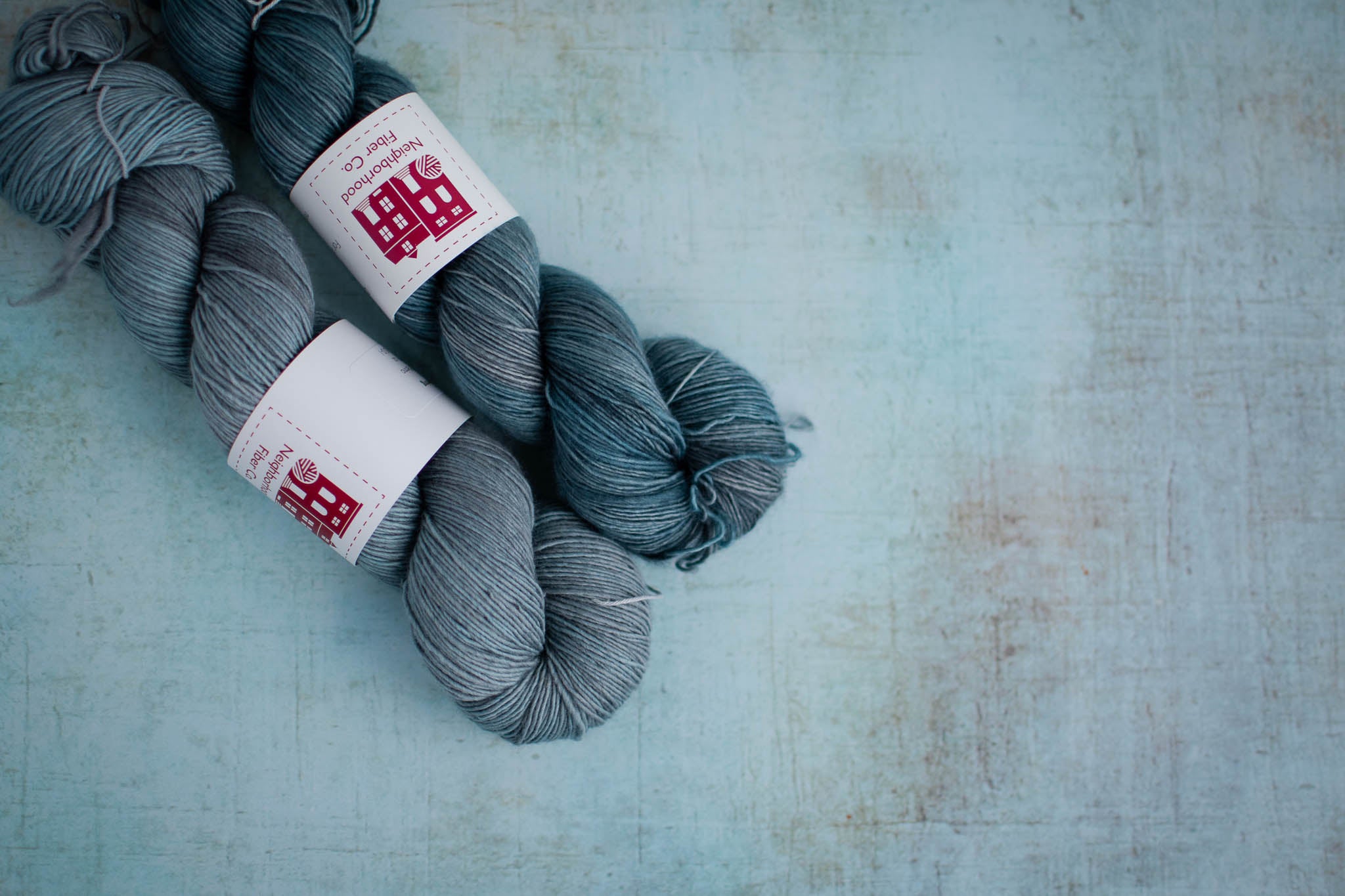 Two skeins of blue grey yarn lie next to each other on a flat surface. The skein to the right is darker and more saturated.