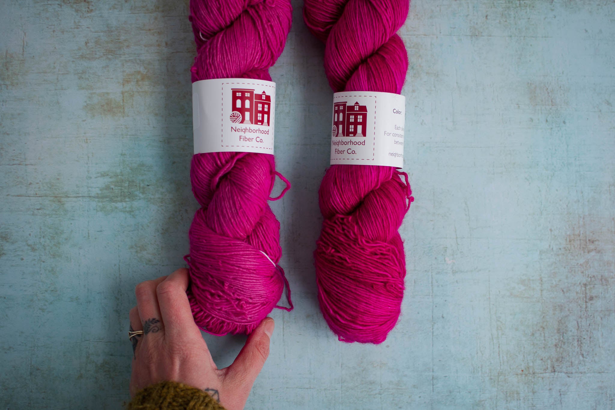 Two skeins of pink yarn, with a hand touching the bottom of the skein on the left. The right skein is darker and more saturated.