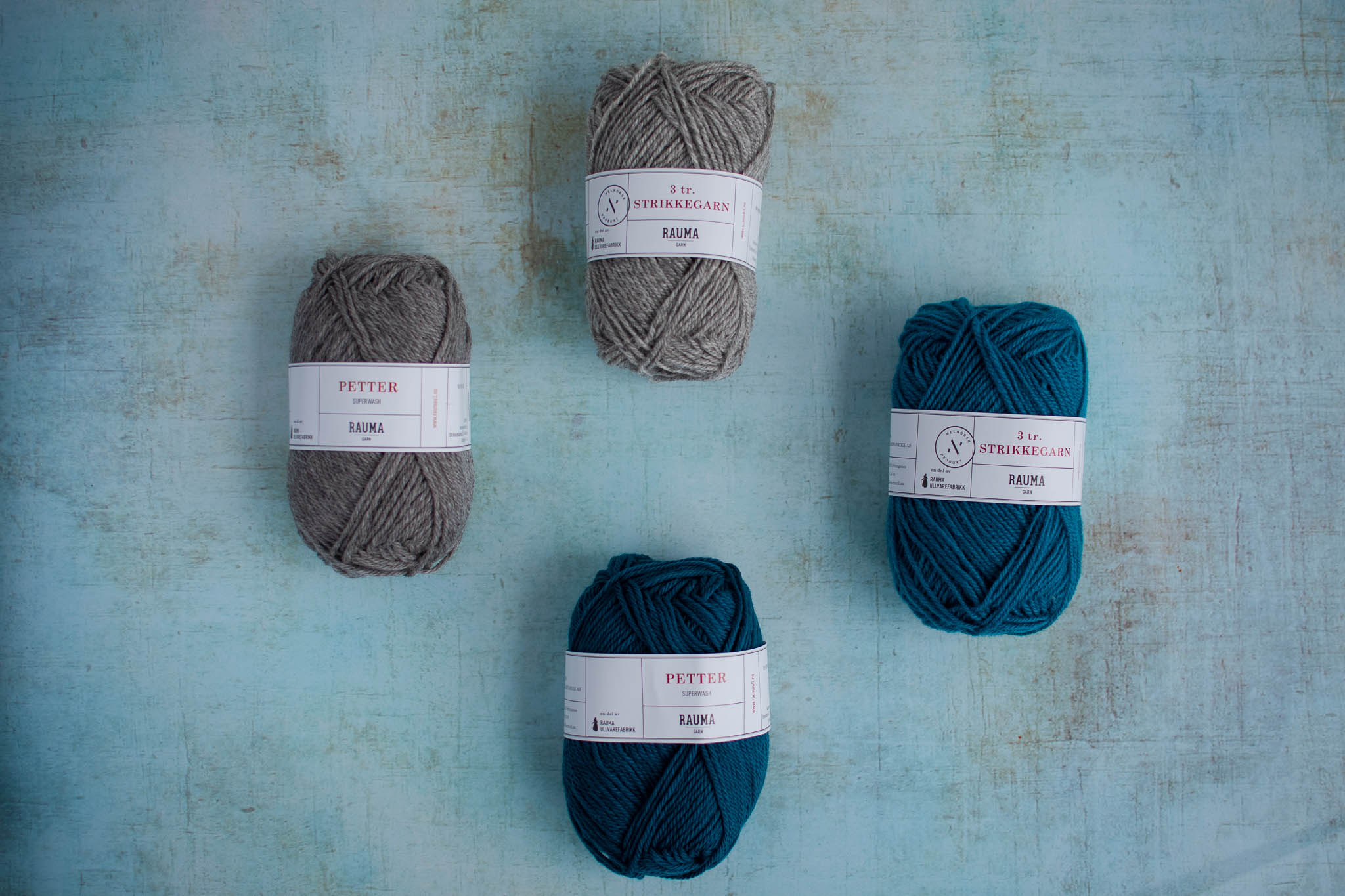 Four balls of yarn arranged on a flat surface, two are grey and two are teal but each of one is slightly darker than the other.