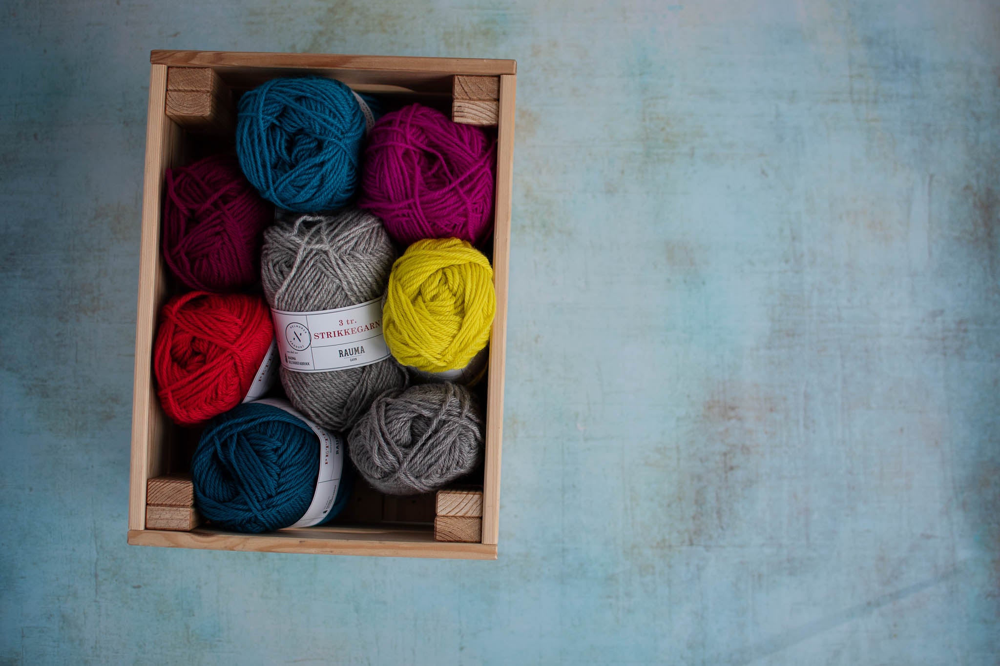 A small wooden box holds balls of yarn in a variety of colours. The box is placed to the left of the image.