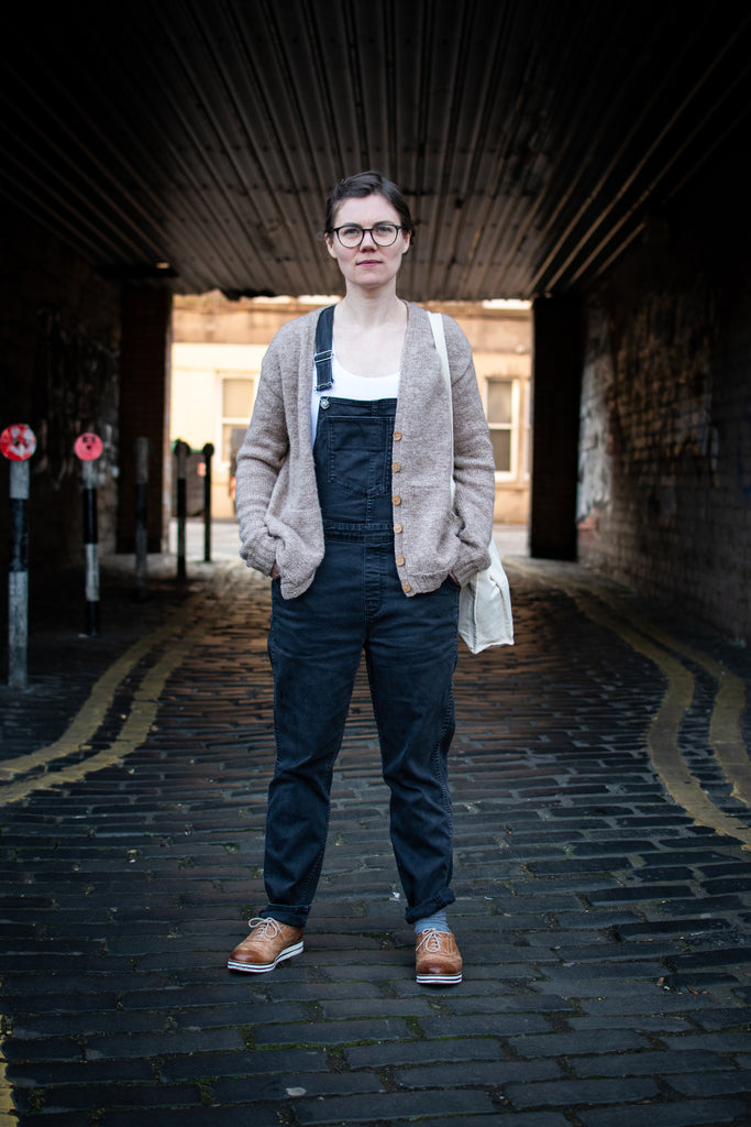 Woman with pale skin wearing dark overalls and a beige wardie cardigan standing with a cobbled alley behind