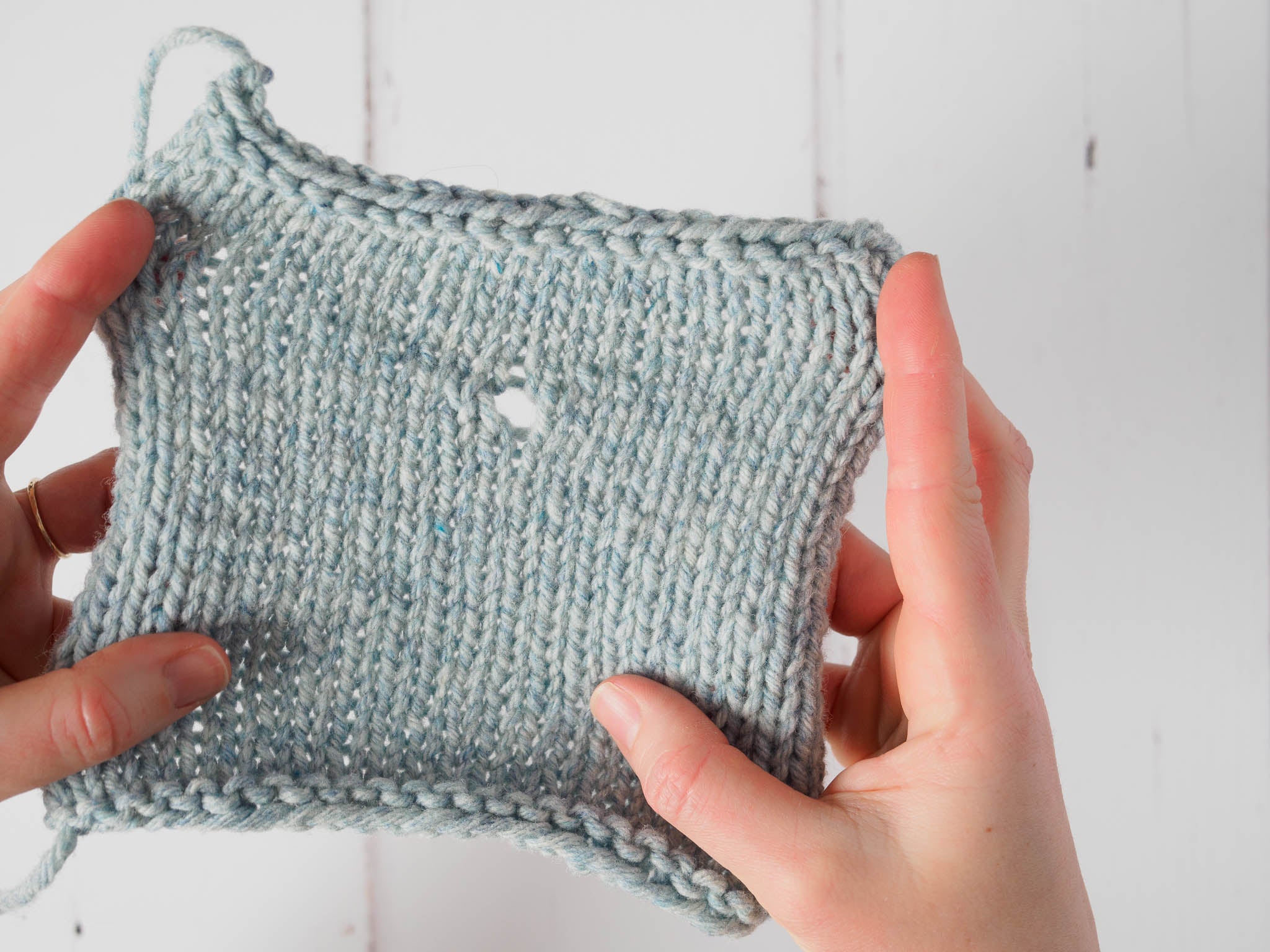 A pale blue swatch with a single yarn over formed in the centre, appearing as a small hole in the fabric. The swatch is held up and being stretched out by a hand on either side.