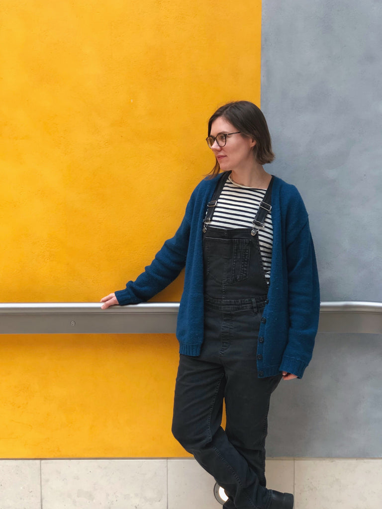 Woman with pale skin wearing dark overalls, a striped shirt and a blue oversized Wardie against a yellow and grey wall