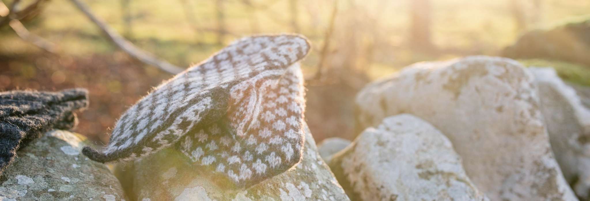 A pair of dark and cream colourwork mittens lie on top of each other, overlapping. They are draped on the top of a stone wall, with trees and a sunset out of focus behind.