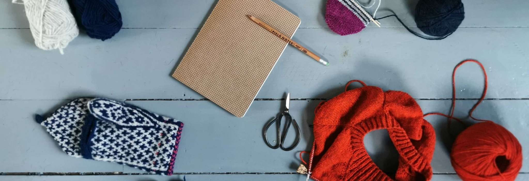 A grey wooden floor with balls of yarn, one unfinished navy and white mitten, a brown notebook and pencil, a small pair of scissors, and an orange sweater in progress next to a ball of orange yarn.