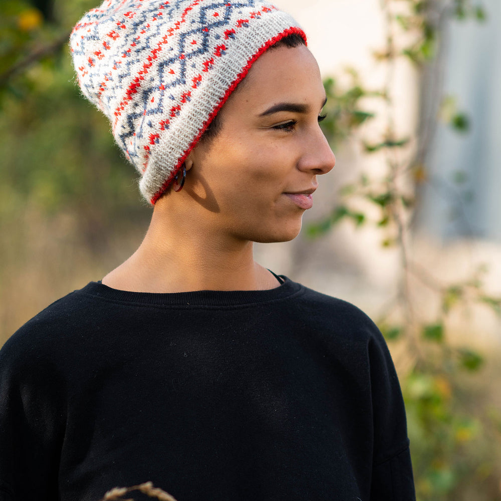 A black woman stands in a wooded area looking towards the right of the frame, they are wearing a black top and a fair isle colourwork hat in a neutral base with red and blue motifs.