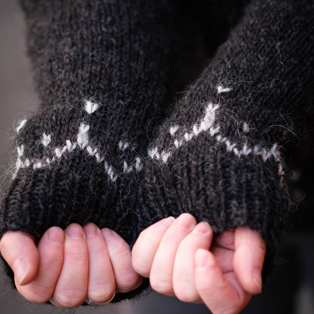 Close up image of white hands with fingers curled in toward the palms, showing off the white colour work detail at the cuffs of black sleeves one a knit sweater.
