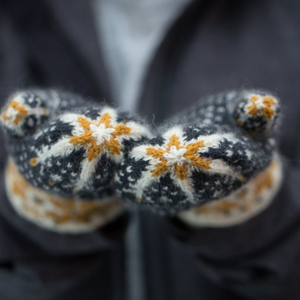 Close up shot of the nordic star detail of the tips of woollen colourwork knit mittens. The stars are gold and white on a navy blue mitten with white flecks.