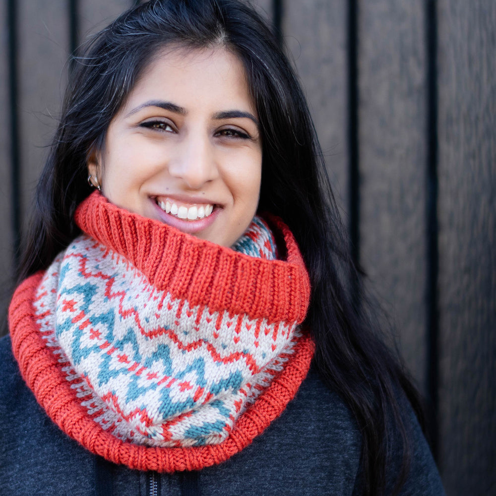 South Asian woman faces the camera smiling, she wears a bright stranded colourwork cowl in coral, teal, and beige. She stands in front of a brown wall