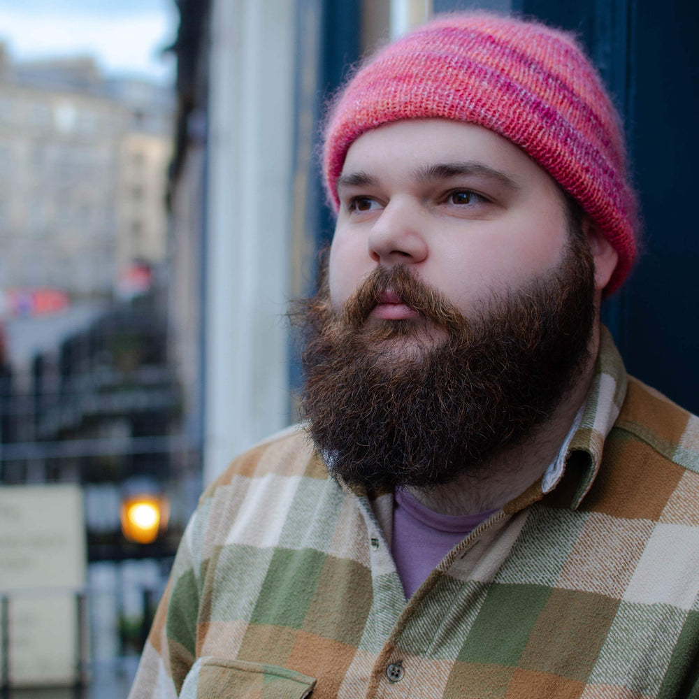 A white man with a bushy brown beard looks up to the right. He is wearing a pink and orange fitted beanie and an orange and green checkered shirt.