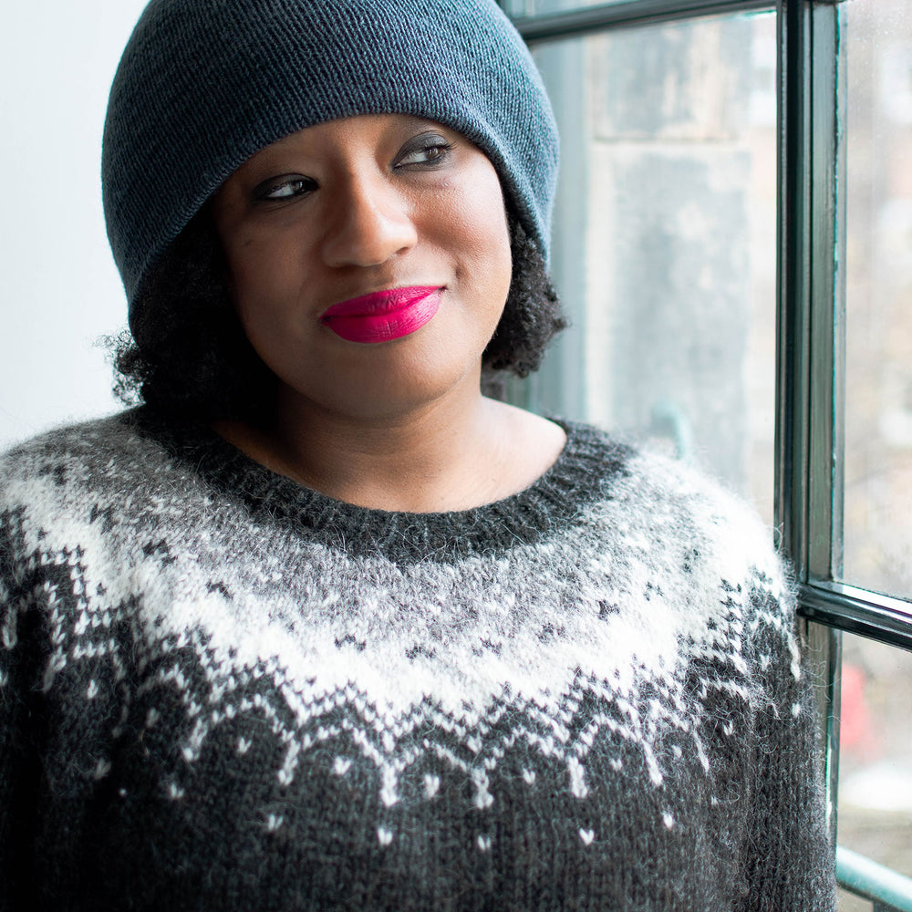 A black woman wearing pink lipstick and a blue beanie stands in front of a window. She&#039;s wearing a black wool jumper with an eye catching white, silver, and grey colourwork yoke.