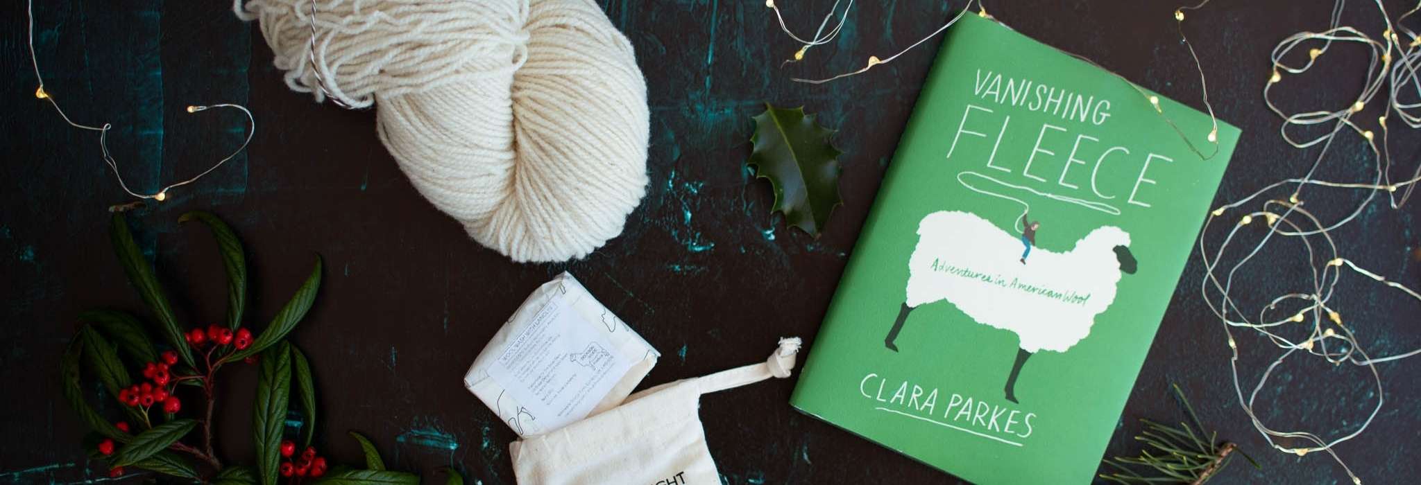 A skein of white yarn, a bar of soap with a cotton drawstring bag and a green book with a drawing of a white sheep on the cover called 'Vanishing Fleece' lie on a dark, flat surface with twinkle lights to the side.