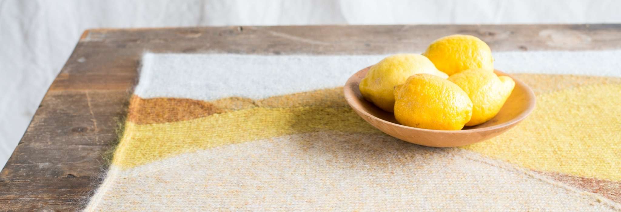 A yellow, orange and white table mat is placed on a wooden table. On top is a small wooden bowl holding four lemons.