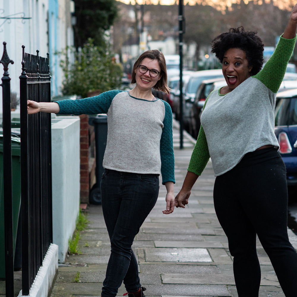 Two women stand joyfully on a city sidewalk back lit by the sunset. Both are wearing black jeans and baseball style kit sweaters. The white woman on the left has glasses and a chin length bob, the sleeves of her top are teal. The black woman on the right has natural hair and the sleeves of her top are green.