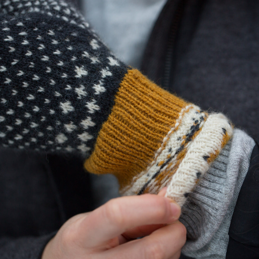 Close up shot of gold rib cuff of woollen colourwork knit mittens that is normally hidden by the decorative white with gold star cuff. The main hand of the mitten is navy blue mitten with white flecks.