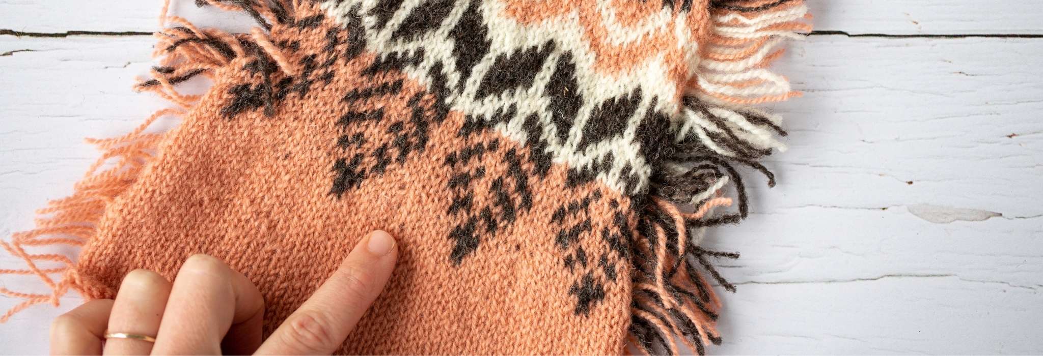 a coral peach, brown and white piece of colourwork knitting, laying on a flat pale surface with fingers of a left hand touching it from the bottom