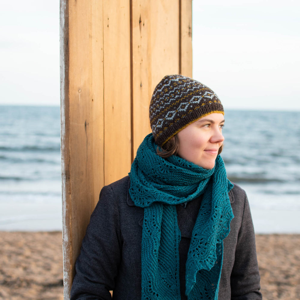 A white woman stands in front of a door on a beach. She&#039;s wearing a grey winter jacket, large turquoise wool shawl wrapped around her neck, and a fair isle beanie. The beanie is a chocolate brown base with light blue and gold motifs.