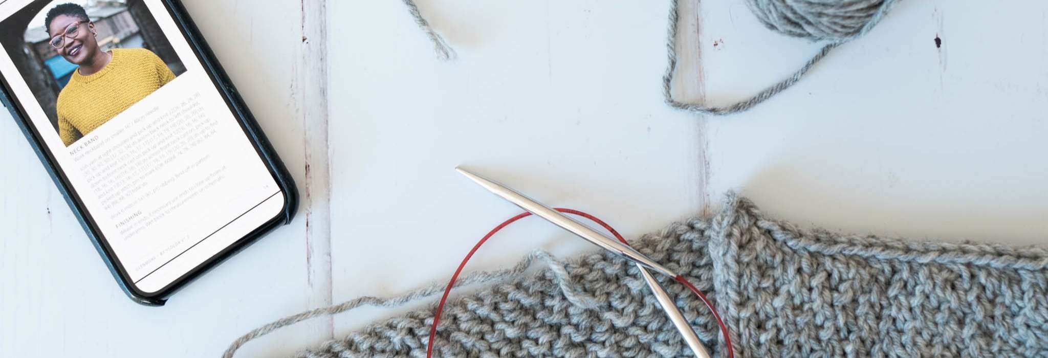 part of a grey sweater laying flat and on knitting needles next to a mobile phone showing an image of the sweater and part of a knitting pattern
