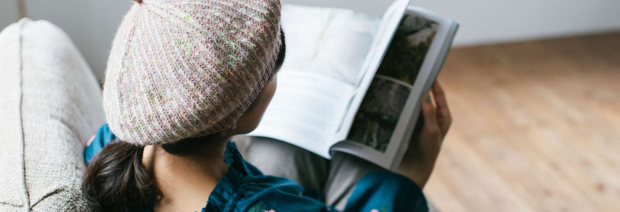 a model wearing a pale grey beret sits holding an open magazine with their back to the camera. They sit on a comfortable grey sofa on a wooden floor.