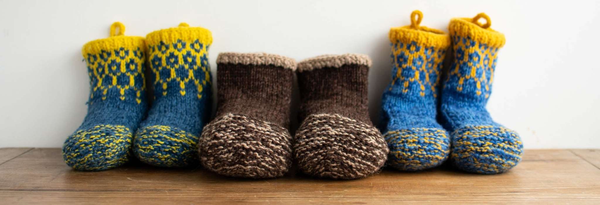 three pairs of knitted slippers lined up against a wall on a wooden surface. The pairs at either end are blue and yellow and the central pair are knitting in shades of brown.