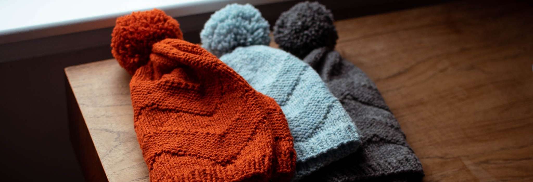 Three beanie hats in orange, blue and grey lie overlapping each other on a wooden table. They have pom poms and a chevron stitch pattern.
