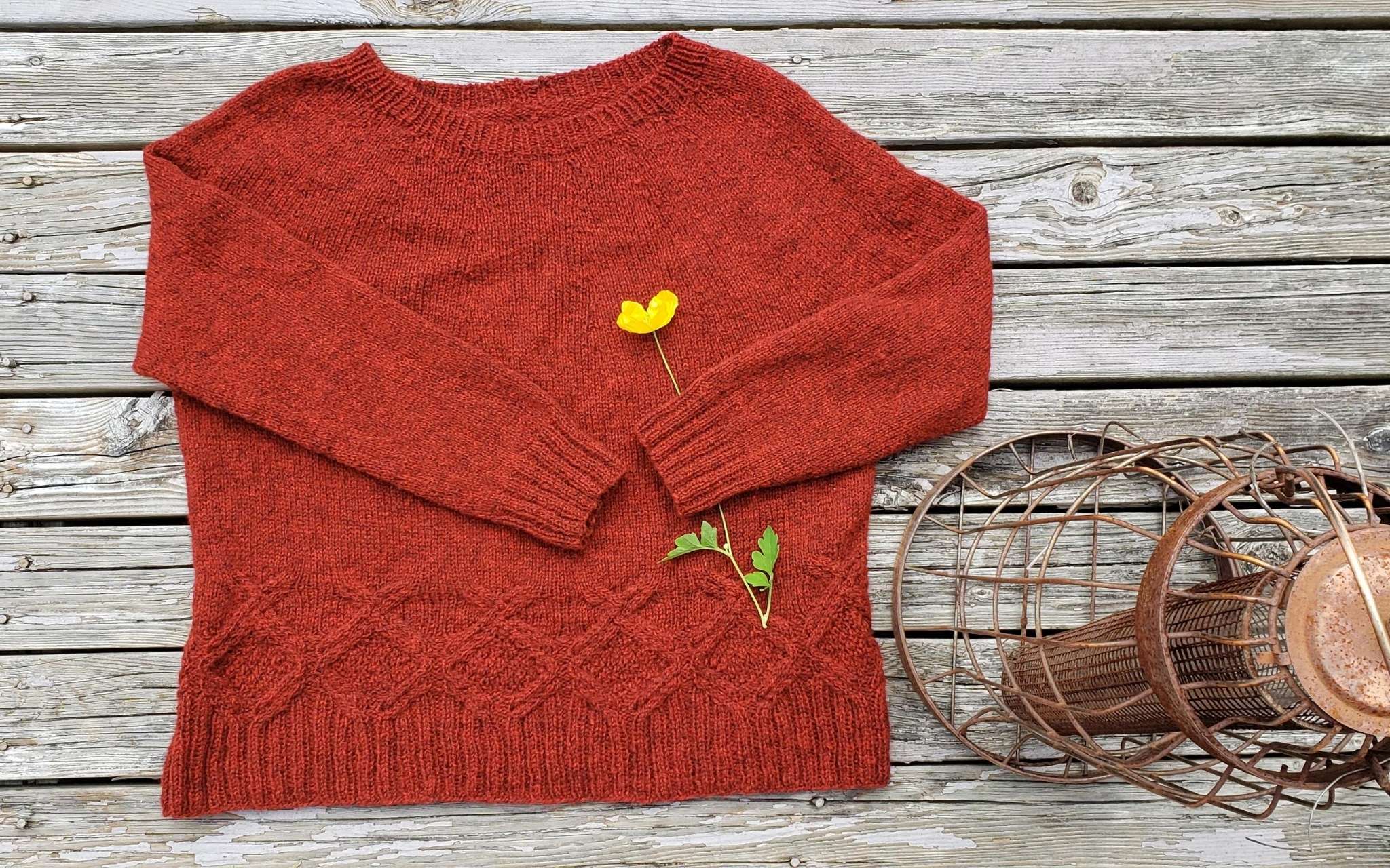 rich orange sweater with cabled hem, laying flat with a yellow flower