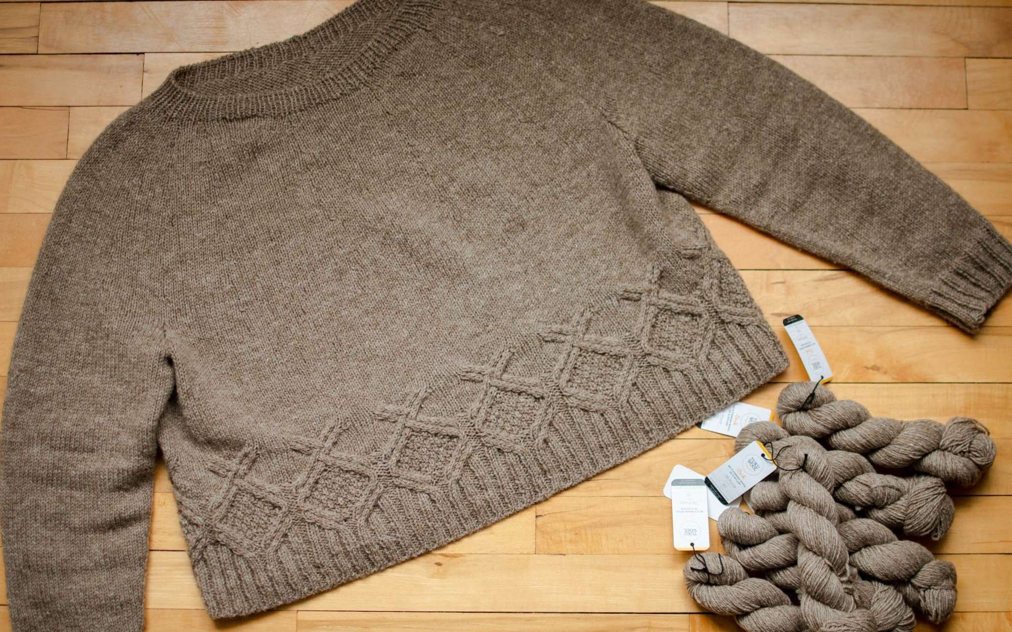 a pale brown neutral sweater with cabled detail around the hem is laid out flat on a wooden surface. Twisted skeins of the same yarn are piled next to it.