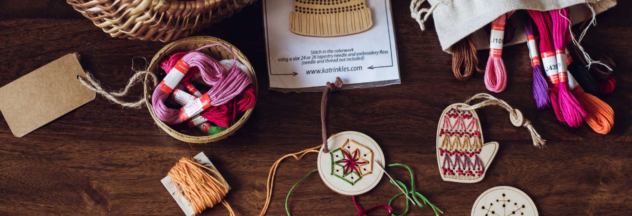 Wooden stitchable ornaments lie on a wooden surface, with stitching done in brightly coloured thread. A pile of threads and a wicker basket lie next to them.
