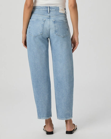 Back view of a person in PAIGE's Alexis Barrel Leg - Verse denim, paired with a white top. The high-rise, light blue jeans feature a barrel leg design and two back pockets. Black sandals complete the look as they stand with legs slightly apart.