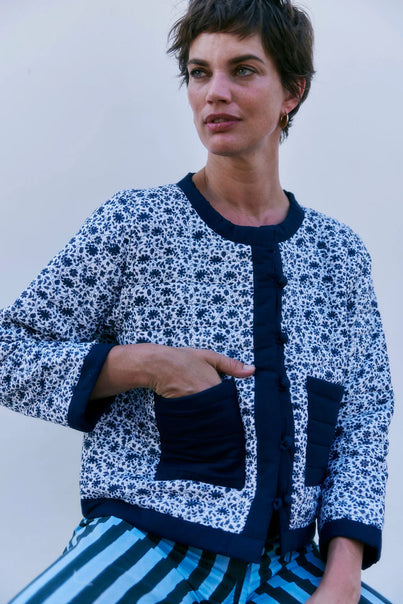 A person stands against a plain background wearing SZ Blockprints' Poppy Reversible Jacket in Navy with patch pockets and a ditsy flower print, paired with striped pants, showcasing a charming ensemble.