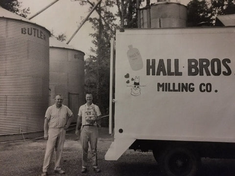 Black and White photograph image of Roy Hall and Hannon Hall, Sr. the founders of Hall's Milling Company standing side by side. Grain bins and a delivery truck are in the background. The words, "Hall Bros. Milling Company" has been painted on the side of the truck.