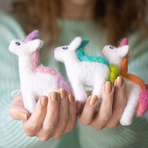 a woman wearing a sea foam sweater holds 3 wool rainbow unicorn ornaments