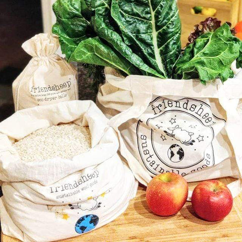 3 cotton Friendsheep bags sit on a wood surface, 2 of the bags hold loose dry rice and green vegetables. 2 red apples sit in front of the bags
