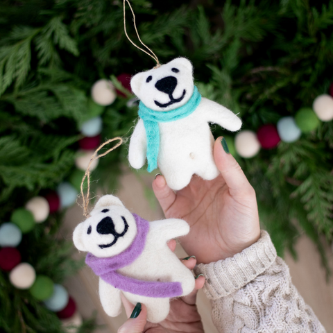 a person holds two smiling white wool polar bear ornaments. The polar bears are wearing a pink and blue scarf and are held in front of green leaves