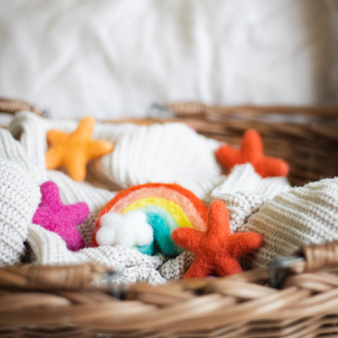 6 vibrant pink and orange wool stars and one wool rainbow ornament lay on a chunky knit blanket in a wicker basket