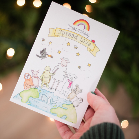 a hand holds a greeting card with a cute drawing of animals holding hands standing on top of the Earth with text "Friendsheep, Spread Love" in front of twinkly lights