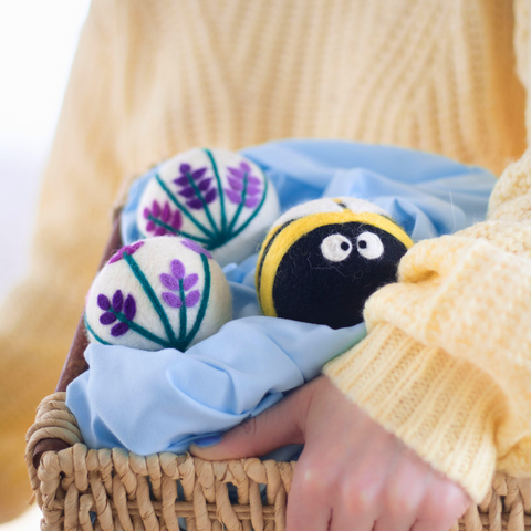 a person wearing a yellow knit sweater holds a woven basket with 3 wool dryer balls (a bee, and 2 white dryer balls with purple and green flowers) laying on blue fabric