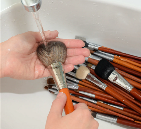 A set of hands washing some makeup brushes in a sink with running water