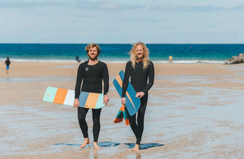 An image of two white male surfers walking up the beach holding wooden bodyboards.