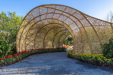 A sculptural structure made from what appears to be bamboo that creates a transparent tunnel through a garden path.