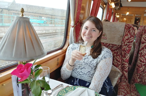 An image of Helen, a young white woman, sitting in a train dining car and toasting the camera with a glass.