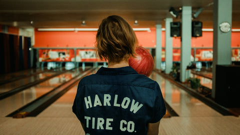 An image of a person wearing a bowling shirt, faced away from the camera, holding a bowling ball and about to bowl.