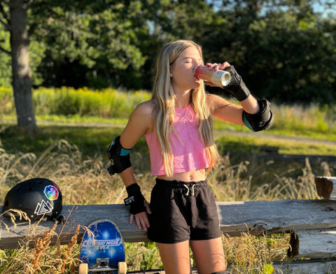 An image of Zoe, a young white teen girl, leaning against a fencepost wearing a pink tank top and shorts, drinking a Wize iced tea.