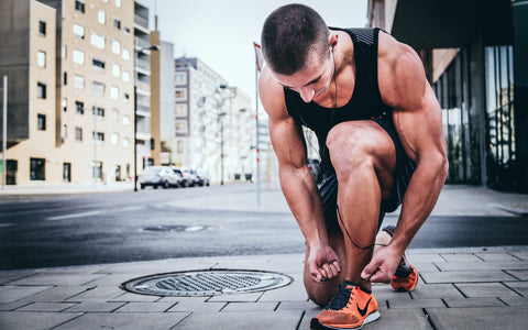 An image of a runner in a black tank top bending down to tie his shoes on a city sidewalk.
