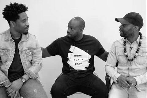 A black and white image of three Black men sitting down, talking to one another. The man in the middle wears a long-sleeve t-shirt that reads "Dope Black Dads"