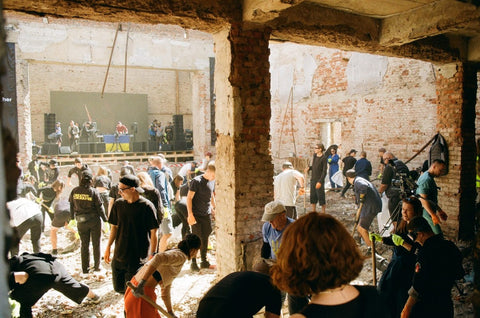 An image of several people cleaning up rubble in a bombed-out building with a DJ spinning records in the background.