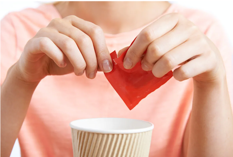 A woman opens an unlabeled sweetener packet over a paper cup.