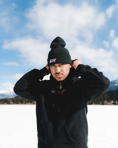 A Black man in a toque against a snowy backdrop pulls up the collar of his black sweater.