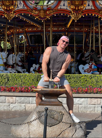 An image of a white man in a grey tank top attempting to pull the sword from the stone at Disneyland.